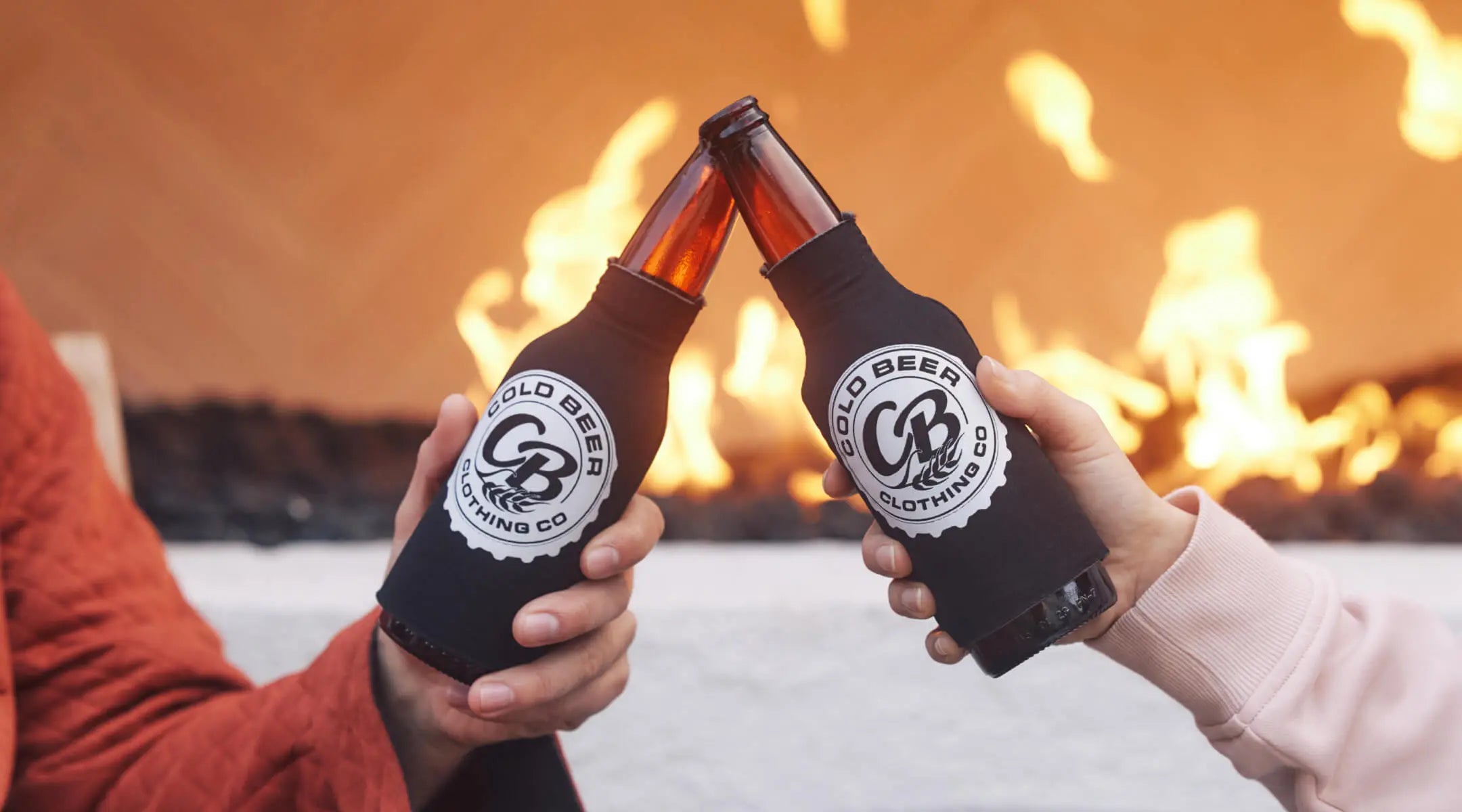 Two beer bottles in Cold Beer Clothing-branded koozies, clinking together in front of a warm fire