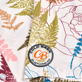 Close-up of the embroidered Cold Beer Clothing Co. logo patch on the left sleeve of the Vintage Big Leaf Button Down, featuring a colorful tropical leaf print.