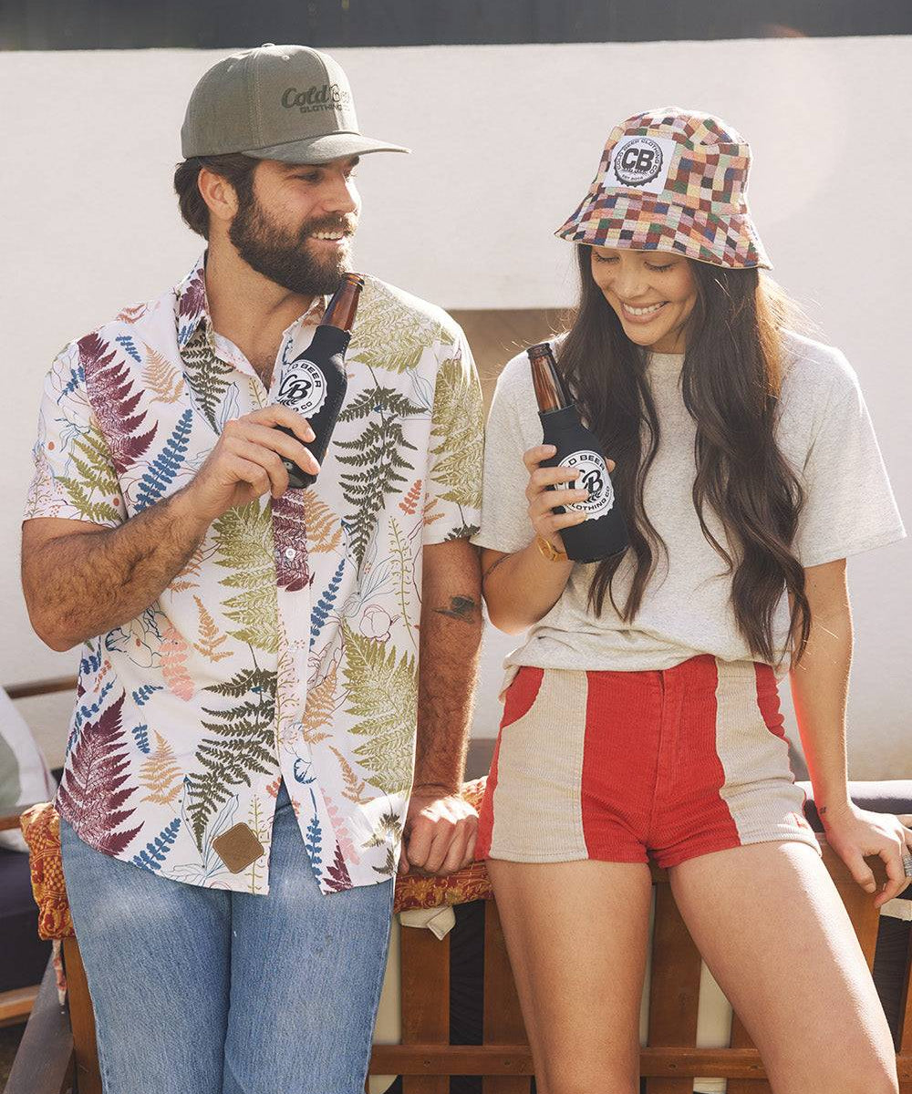 Man wearing the Vintage Big Leaf Button Down by Cold Beer Clothing Co., featuring a colorful tropical leaf print, standing with a woman in CBC apparel while enjoying cold beers.