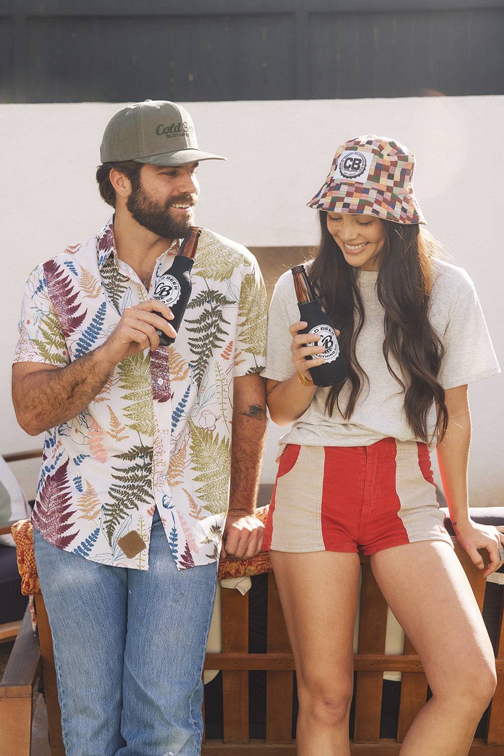 Man wearing the Vintage Big Leaf Button Down by Cold Beer Clothing Co., featuring a colorful tropical leaf print, standing with a woman in CBC apparel while enjoying cold beers.